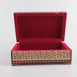 May include: A rectangular wooden box with an open lid, featuring a vibrant red velvet interior lining. The exterior sides are decorated with a detailed geometric pattern in shades of black, cream, and gold, finished with a small star border.
