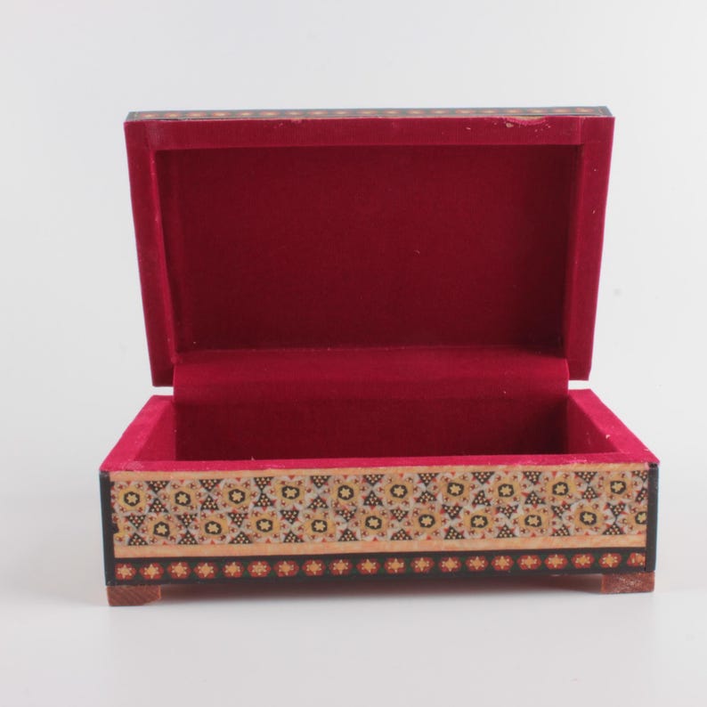 May include: A rectangular wooden box with an open lid, featuring a vibrant red velvet interior lining. The exterior sides are decorated with a detailed geometric pattern in shades of black, cream, and gold, finished with a small star border.