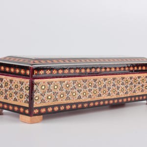 May include: A rectangular wooden box with intricate inlaid geometric patterns in shades of brown, gold, and white. The box has a hinged lid and sits on four small feet.