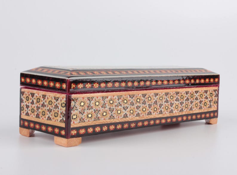 May include: A rectangular wooden box with intricate inlaid geometric patterns in shades of brown, gold, and white. The box has a hinged lid and sits on four small feet.