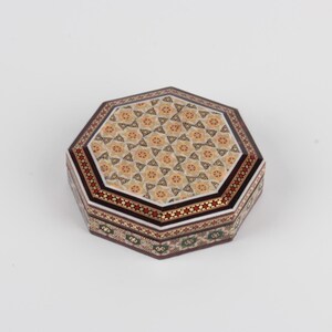 May include: An octagonal decorative box with a detailed mosaic design. The lid features a repeating floral pattern in beige and gold, framed by a border of red and white stars. The sides showcase intricate geometric patterns in green, red, and gold.