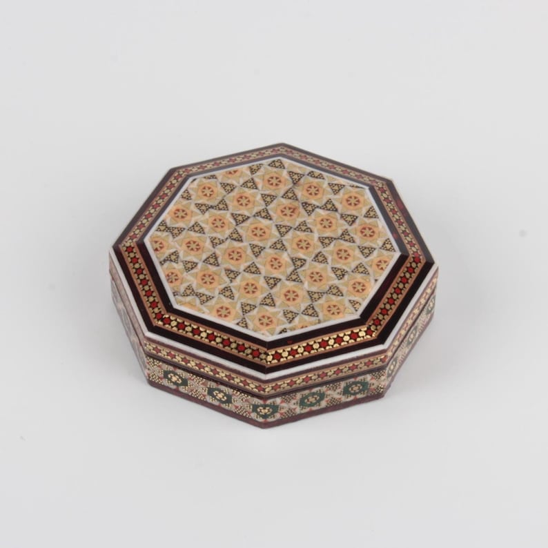 May include: An octagonal decorative box with a detailed mosaic design. The lid features a repeating floral pattern in beige and gold, framed by a border of red and white stars. The sides showcase intricate geometric patterns in green, red, and gold.