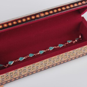 May include: A turquoise and copper bracelet with a teardrop design sits inside a small, ornate wooden box with a red velvet interior. The box is decorated with intricate floral patterns.