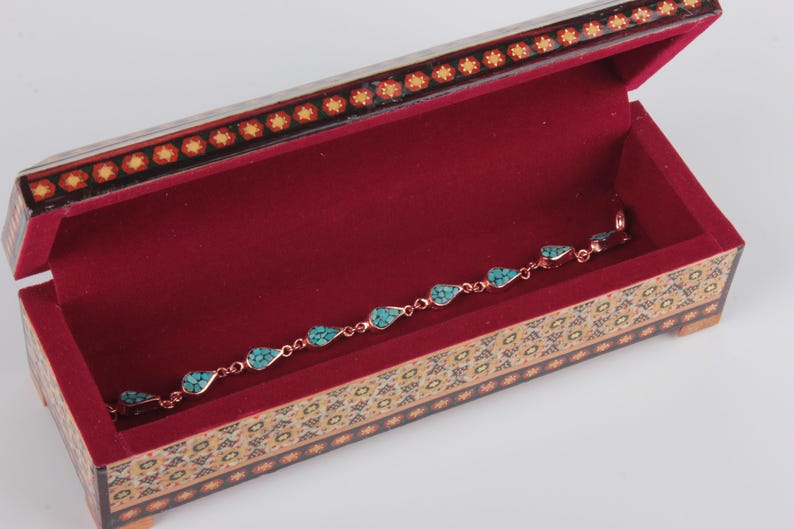 May include: A turquoise and copper bracelet with a teardrop design sits inside a small, ornate wooden box with a red velvet interior. The box is decorated with intricate floral patterns.