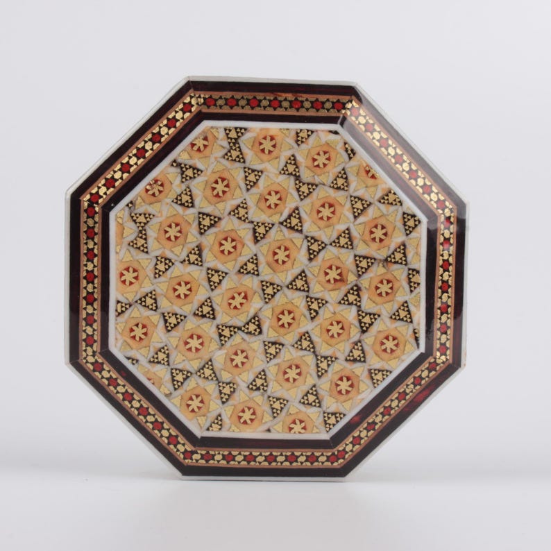 May include: An octagonal decorative box with an intricate geometric pattern. The box features a central design of repeating star-like shapes in shades of yellow and brown. The border is a dark brown with red and gold accents.