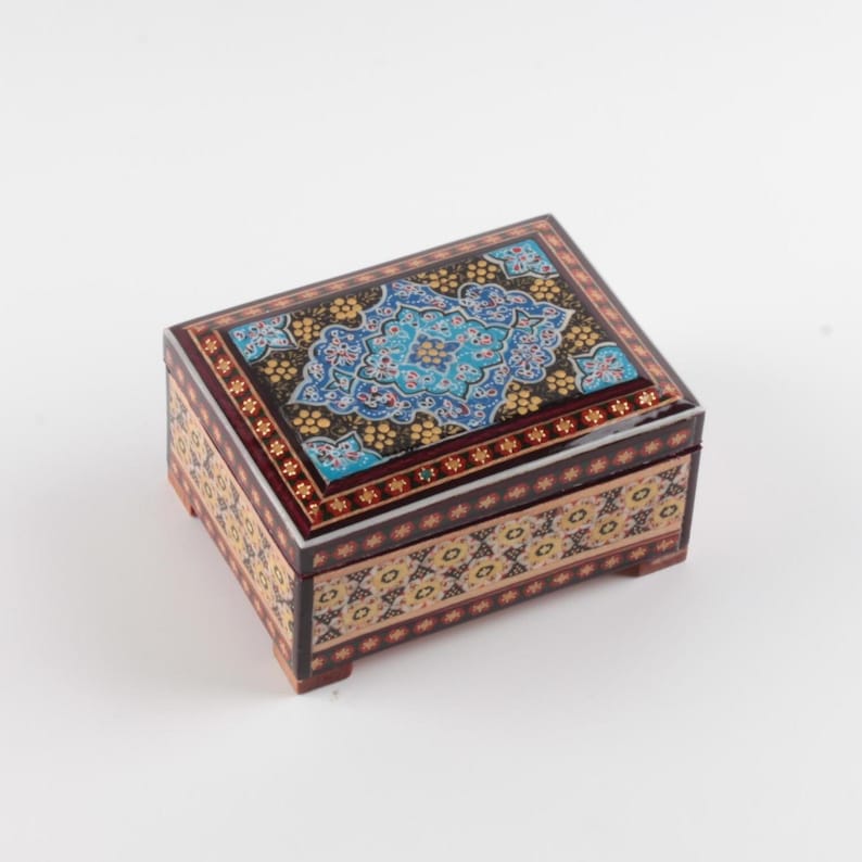 May include: A rectangular wooden box featuring a lid with a central blue and gold floral pattern on a black background. The sides of the box are decorated with a repeating geometric pattern in shades of cream, black, and gold. The box has four small feet.