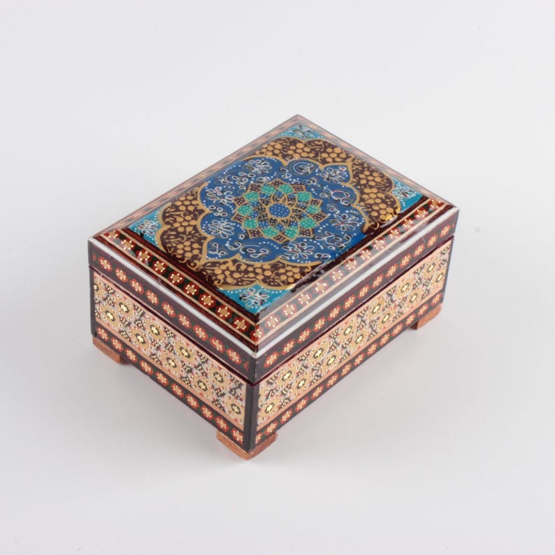 May include: A rectangular wooden box featuring intricate hand-painted floral patterns in shades of blue, gold, and black. The lid displays a central mandala-like design, while the sides are decorated with repeating geometric motifs and four small feet.
