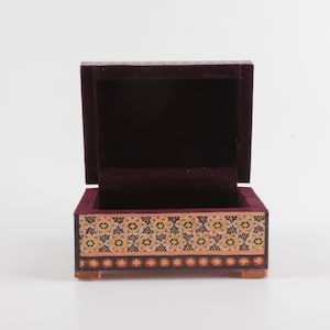May include: A small rectangular wooden box with an open lid, featuring a dark purple velvet interior lining. The exterior front panel displays a decorative pattern of repeating floral motifs in shades of yellow, white, and black on a tan background.