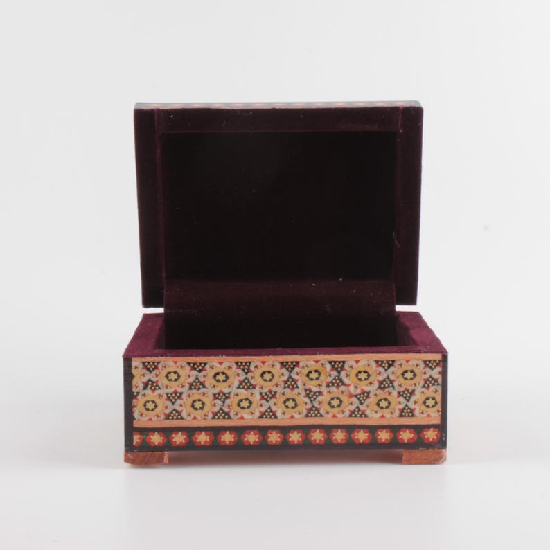 May include: A small rectangular wooden box with an open lid, featuring a dark purple velvet interior lining. The exterior front panel displays a decorative pattern of repeating floral motifs in shades of yellow, white, and black on a tan background.