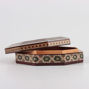 May include: An open, hexagonal wooden box with a decorative lid. The lid and base are adorned with intricate patterns of red, gold, and green floral designs. The box is set against a white background.