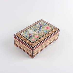 May include: A rectangular wooden box featuring a hand-painted lid with two peacocks surrounded by pink flowers and green foliage. The sides of the box are decorated with intricate black, gold, and white geometric patterns and small feet.