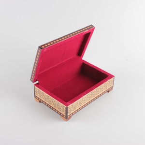 May include: A rectangular wooden box with an open lid, featuring intricate geometric inlay patterns on the exterior sides and edges. The interior of the box is lined with a vibrant red velvet fabric, and the base is supported by four small feet.