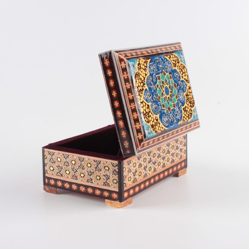 May include: A rectangular wooden box with an open lid, featuring intricate geometric marquetry patterns in shades of brown, cream, and black. The interior is lined with dark maroon velvet, and the lid displays a vibrant blue and gold floral design.