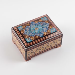 May include: A rectangular wooden box with a hinged lid, featuring intricate hand-painted floral patterns in shades of blue, gold, and brown. The exterior displays a dark brown base with repeating geometric and botanical motifs on all visible sides.