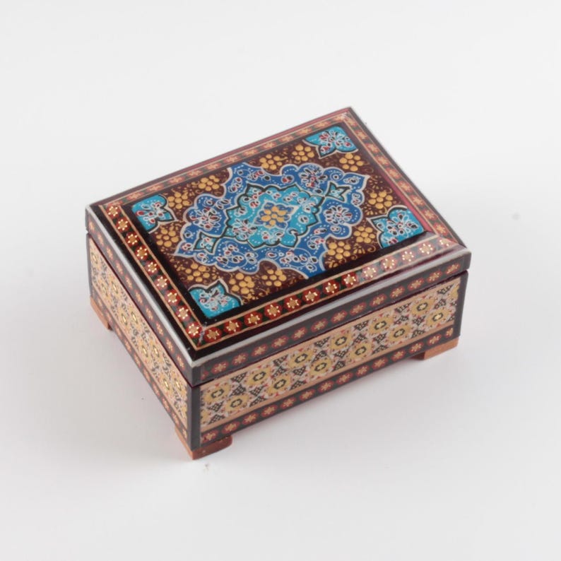 May include: A rectangular wooden box with a hinged lid, featuring intricate hand-painted floral patterns in shades of blue, gold, and brown. The exterior displays a dark brown base with repeating geometric and botanical motifs on all visible sides.