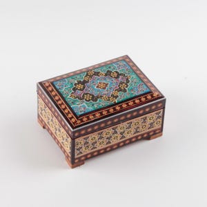 May include: A rectangular wooden box featuring intricate marquetry and inlaid patterns. The lid displays a vibrant turquoise, black, and gold floral design, while the sides are decorated with a repeating geometric motif in cream, black, and brown.