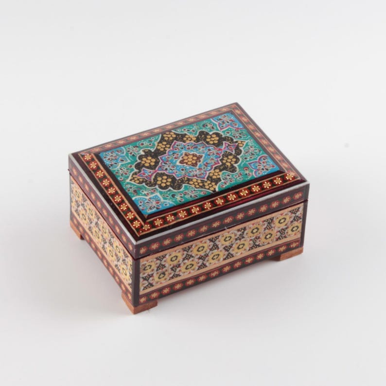 May include: A rectangular wooden box featuring intricate marquetry and inlaid patterns. The lid displays a vibrant turquoise, black, and gold floral design, while the sides are decorated with a repeating geometric motif in cream, black, and brown.