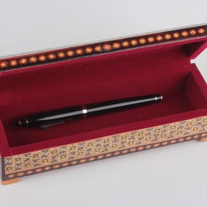 May include: A small, rectangular wooden box with a red velvet interior and intricate floral patterns on the exterior. The box is open and a black pen is lying inside.