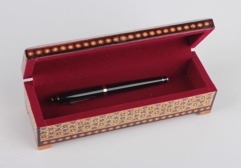 May include: A small, rectangular wooden box with a red velvet interior and intricate floral patterns on the exterior. The box is open and a black pen is lying inside.