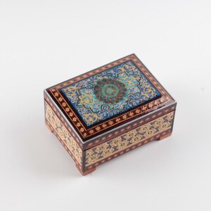 May include: A rectangular wooden box featuring a lid with a central blue, gold, and black floral medallion pattern. The sides of the box are decorated with a repeating cream and black geometric pattern, accented by small red and gold flower motifs.