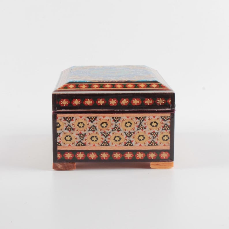 May include: A rectangular wooden box with a hinged lid, featuring intricate geometric patterns in shades of red, black, white, and tan. The box has a dark brown finish, a flat top, and four small, light-colored wooden feet at the base corners.