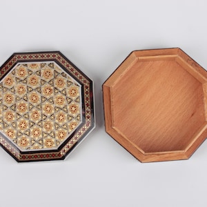 May include: An open, octagonal wooden box with a decorative lid. The lid features a detailed mosaic pattern in shades of brown, beige, and black. The box's interior is a light brown color, and the box is set against a white background.