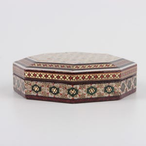 May include: An octagonal decorative box with a detailed mosaic design. The box features a dark brown base with intricate patterns in red, gold, and green. The lid has a cream-colored mosaic pattern with a border of red and gold.