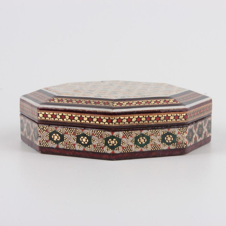 May include: An octagonal decorative box with a detailed mosaic design. The box features a dark brown base with intricate patterns in red, gold, and green. The lid has a cream-colored mosaic pattern with a border of red and gold.