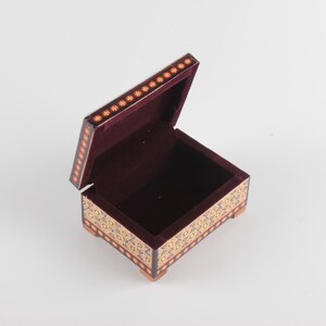 May include: A small rectangular wooden box with an open hinged lid. The exterior features a dark brown finish with intricate gold and black floral patterns along the sides and a row of small orange flower motifs on the lid edge. The interior is lined.