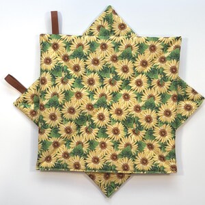 May include: Two square quilted pot holders featuring a repeating pattern of yellow sunflowers with brown centers and green leaves on a light background. Each pot holder includes a small brown fabric loop attached to one corner for hanging.