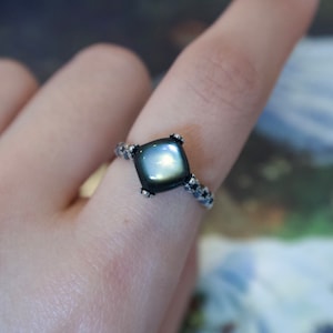 May include: A silver ring with a square, iridescent gemstone. The ring's band has a delicate, textured design with small, dark accents. The gemstone reflects a range of colours, creating a captivating visual effect. This piece of jewellery is a statement accessory.