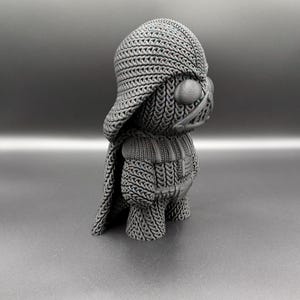 May include: A gray, 3D-printed figurine of Darth Vader, featuring a knitted texture. The figure has a helmet, cape, and chest armor, all rendered in a detailed, textured pattern. The overall design is a stylized representation of the iconic character.