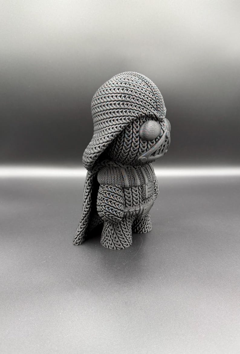 May include: A gray, 3D-printed figurine of Darth Vader, featuring a knitted texture. The figure has a helmet, cape, and chest armor, all rendered in a detailed, textured pattern. The overall design is a stylized representation of the iconic character.
