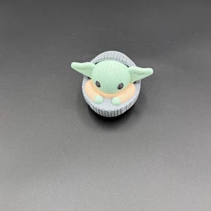 May include: A green and tan character figurine in a gray knitted carrier. The figurine has large ears and black eyes. The carrier is oval-shaped and has a knitted texture. The background is a solid gray color.