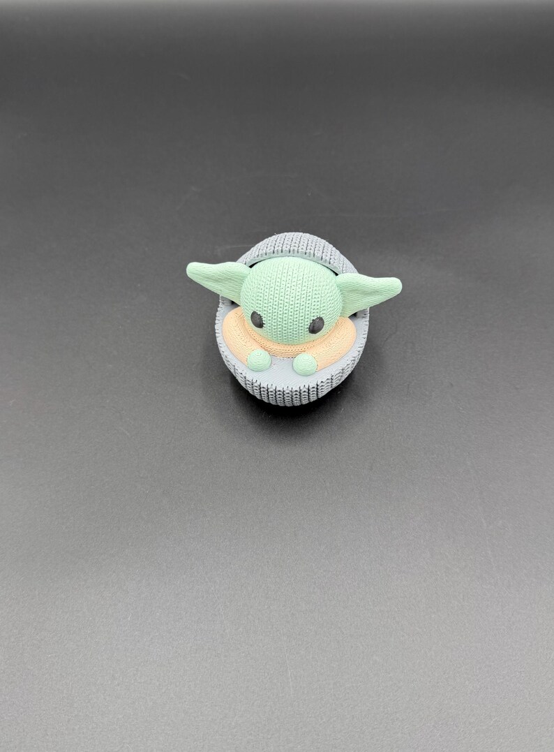 May include: A green and tan character figurine in a gray knitted carrier. The figurine has large ears and black eyes. The carrier is oval-shaped and has a knitted texture. The background is a solid gray color.