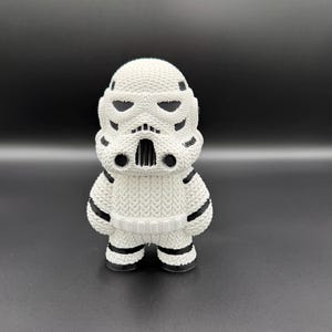May include: A white and black Stormtrooper figurine. The figurine has a knitted texture, with black accents on the helmet, shoulders, and legs. The figure stands upright on a dark surface.