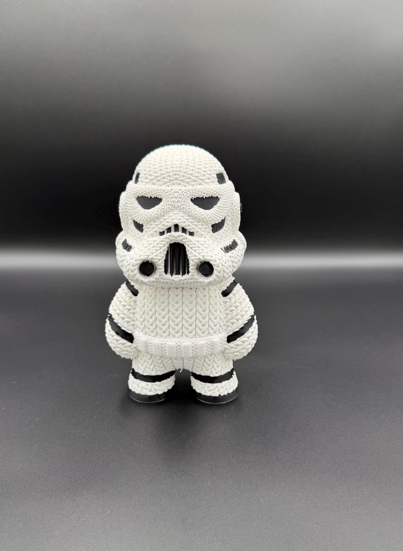 May include: A white and black Stormtrooper figurine. The figurine has a knitted texture, with black accents on the helmet, shoulders, and legs. The figure stands upright on a dark surface.