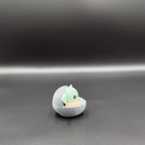 May include: A small, knitted figurine of a green alien-like character resting in a gray, spherical cradle. The cradle has a textured, woven appearance. The character has large eyes and pointed ears. The background is a gradient of dark gray.