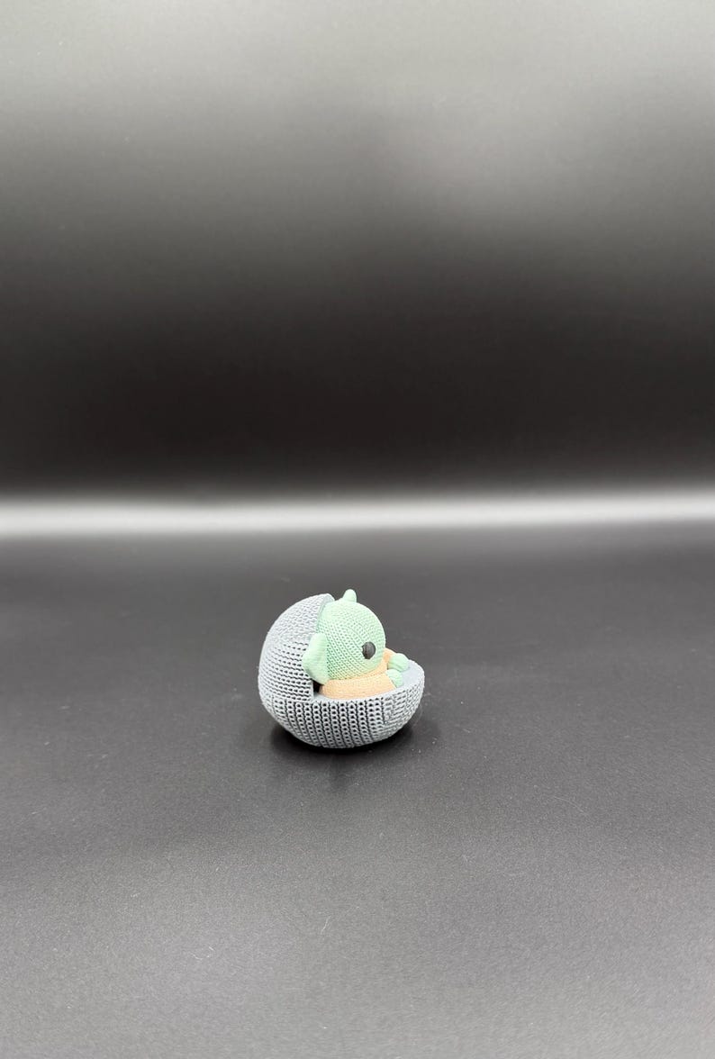 May include: A small, knitted figurine of a green alien-like character resting in a gray, spherical cradle. The cradle has a textured, woven appearance. The character has large eyes and pointed ears. The background is a gradient of dark gray.