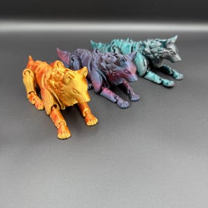 May include: Three articulated wolf figurines in shades of orange, purple, and teal. The wolves are lying down, with their legs extended. The figurines have detailed fur textures and are made of a plastic material.