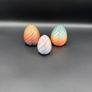 May include: Three decorative, egg-shaped objects with a gradient color scheme. The eggs range in size and feature a striped design. Colors include orange, red, blue, and white. The objects are set against a dark background.