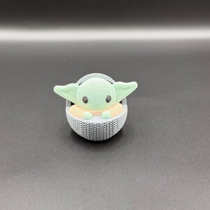 May include: A 3D-printed figurine of a character with large ears and big eyes, seated in a woven-style carrier. The character has a green head and ears, with a tan body. The carrier is gray.