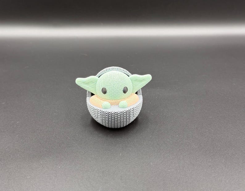 May include: A 3D-printed figurine of a character with large ears and big eyes, seated in a woven-style carrier. The character has a green head and ears, with a tan body. The carrier is gray.