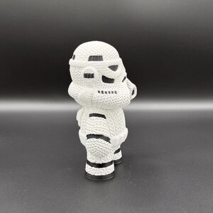 May include: A white, knitted Stormtrooper figurine. The figure has black accents on the helmet and body, with a textured appearance. The figurine is standing on a dark surface.