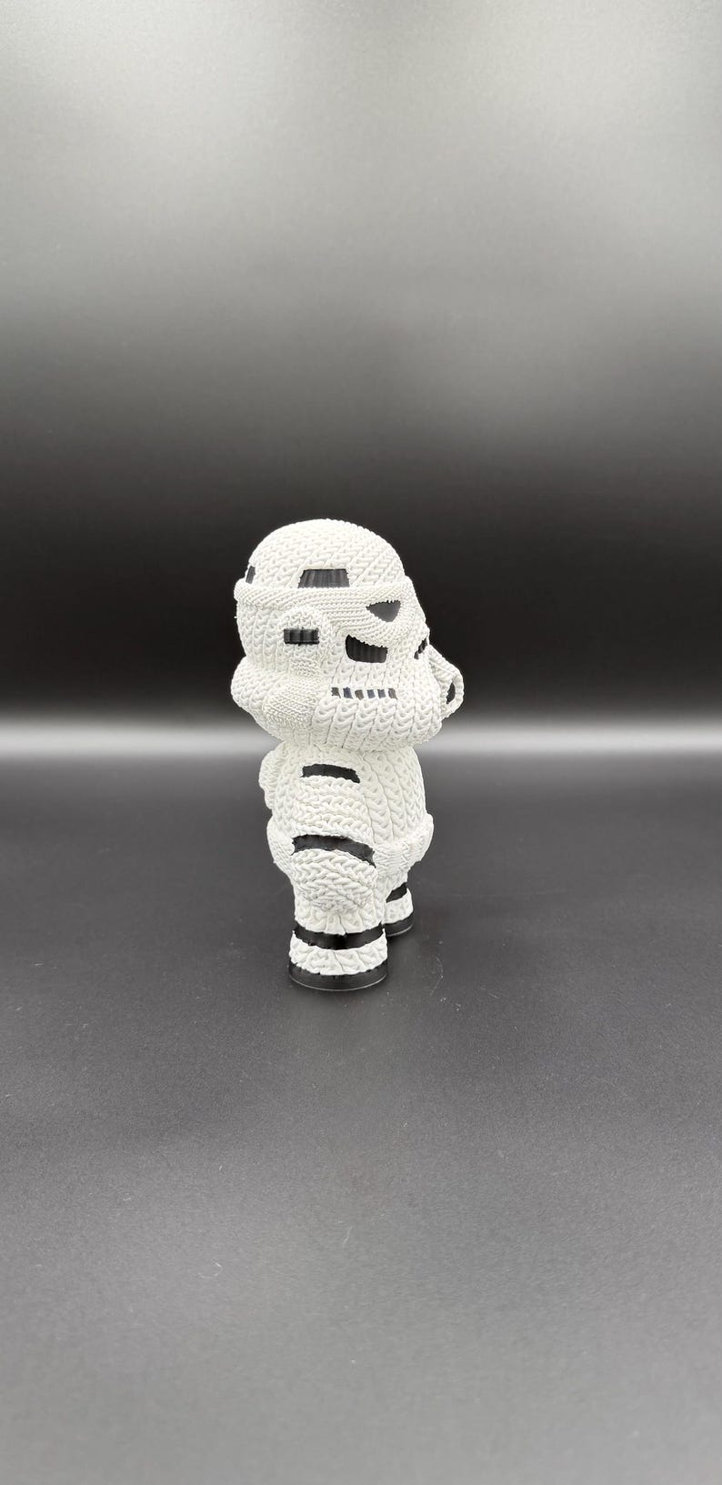 May include: A white, knitted Stormtrooper figurine. The figure has black accents on the helmet and body, with a textured appearance. The figurine is standing on a dark surface.