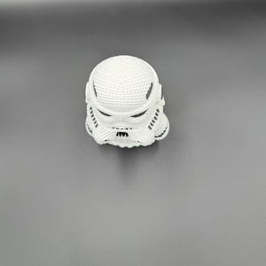 May include: A white Stormtrooper helmet figurine with black accents. The helmet has a textured surface and is set against a gray background. The design is a recognizable element from the Star Wars franchise.