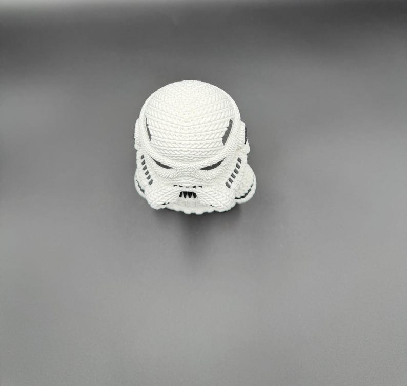 May include: A white Stormtrooper helmet figurine with black accents. The helmet has a textured surface and is set against a gray background. The design is a recognizable element from the Star Wars franchise.