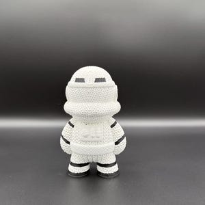 May include: A white figurine of a character in a knitted style, with black accents. The figure has a rounded head, a cylindrical body, and short legs. The design includes black stripes on the arms and legs, and black rectangular eye shapes.