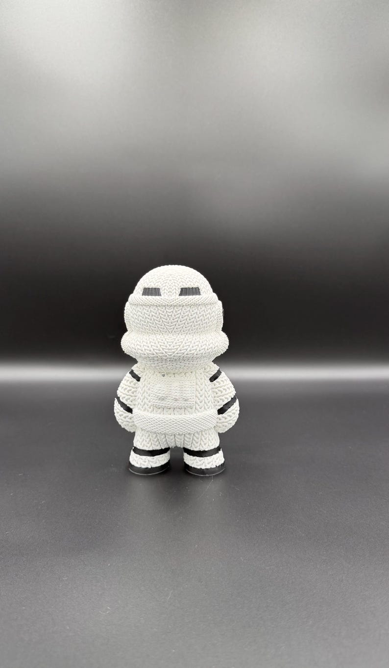 May include: A white figurine of a character in a knitted style, with black accents. The figure has a rounded head, a cylindrical body, and short legs. The design includes black stripes on the arms and legs, and black rectangular eye shapes.