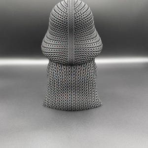 May include: A gray, textured sculpture of a figure with a rounded head and a flowing, skirt-like base. The sculpture has a woven or chainmail-like appearance, with intricate patterns across its surface. The figure is set against a dark gray background.